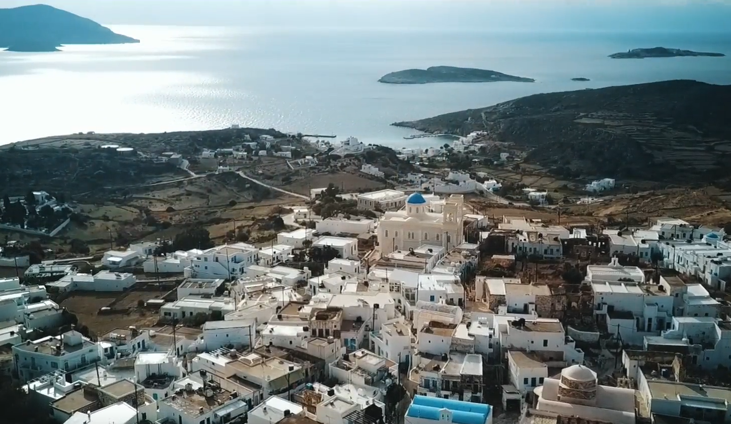 Εναέριο πλάνο από τη Κίμωλο με πολλά άσπρα κυκλαδίτικα σπίτια, μία εκκλησία και τη θάλασσα στο βάθος.