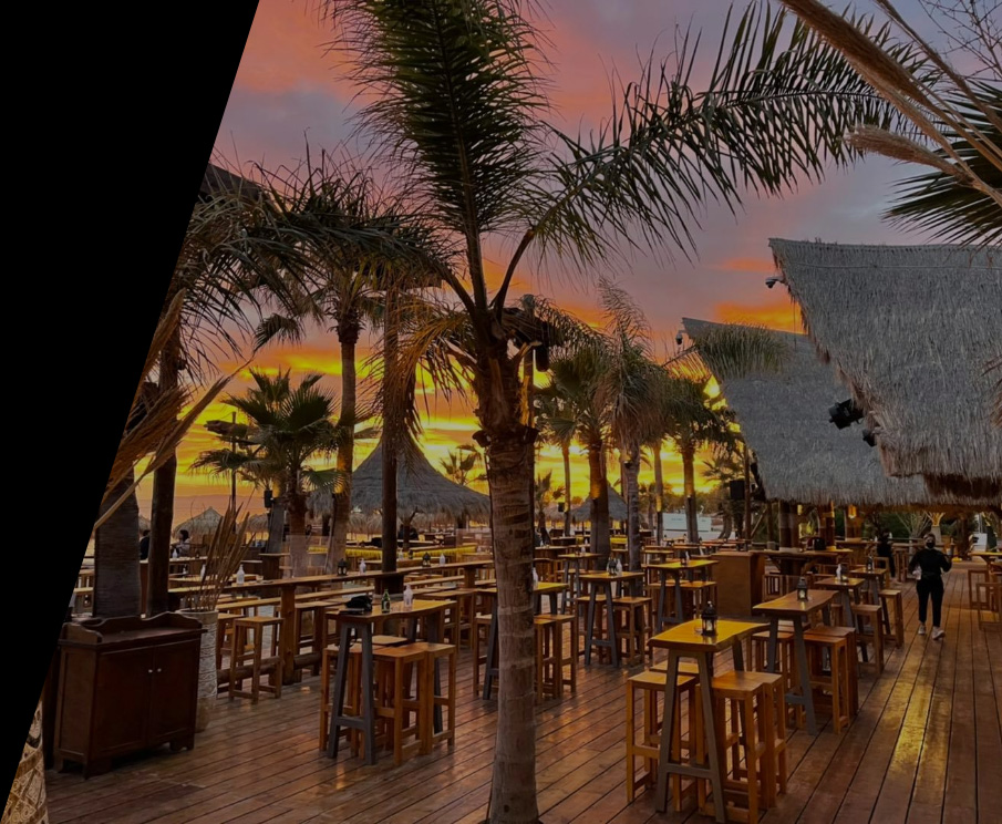 Το Bolivar Beach Bar την ώρα του ηλιοβασιλέματος. Υπάρχουν ψηλά ξύλινα τραπέζια και ξύλινα σκαμπό σε όλο τον χώρο και ανάμεσά τους φοίνικες. 
