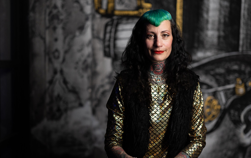 Πορτραίτο της Electric Mama, της πρώτης Αθηναίας tattoo artist με δικό της tattoo studio. Βρίσκεται μπροστά από τοίχο με ένα σκούρο γκρι γκράφιτι. Έχει μαύρα μακριά μαλλιά, ενώ οι αφέλειές της έχουν έντονο πράσινο χρώμα. Φορά χρυσή μεταλλιζέ μπλούζα και μαύρο γιλέκο, κόκκινο κραγιόν, και ο λαιμός της είναι καλυμμένος με τατουάζ σε κόκκινο χρώμα.

